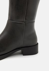 Zwarte leren laarzen tot de knie met een gladde textuur, vierkante neus en lage blokhak. Heeft subtiele stiksels langs de naad.