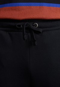 Sorte joggingbukser med snor i taljen, udstyret med en sølvøje og en glat tekstur. Baggrunden har en rød og blå stribet top.