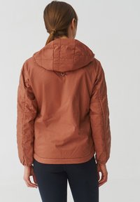 Veste légère et rembourrée de couleur rouille, dotée d'une capuche et d'un design matelassé. Texture lisse avec des poignets élastiques et un ourlet ajusté.