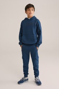 Mornarško modra kapuca z napisom "BLUE", kombinirana s usklajenimi cargo hlačami. Obe stvari imata mehak otip in elastične manšete. Tenisice dopolnjujejo videz.