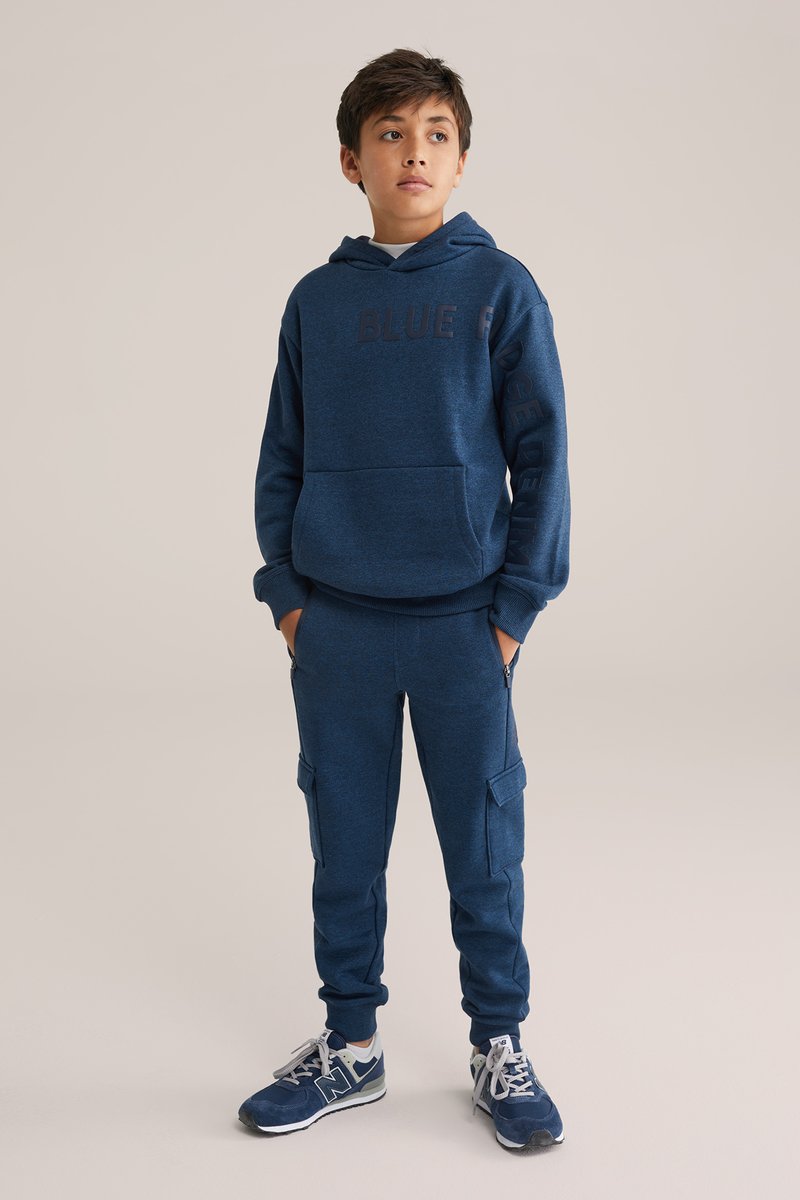 Mornarško modra kapuca z napisom "BLUE", kombinirana s usklajenimi cargo hlačami. Obe stvari imata mehak otip in elastične manšete. Tenisice dopolnjujejo videz.