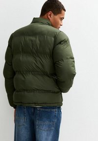 Giacca a vento verde oliva con colletto alto, caratterizzata da una texture trapuntata e una vestibilità comoda. Indossata sopra pantaloni in denim con tasche posteriori.