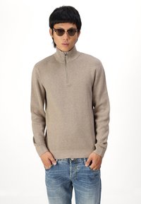 Beige zip-up sweater met een hoge kraag, gemaakt van een zacht materiaal. Gekocht met lichtblauwe denim jeans. Model draagt een zonnebril.