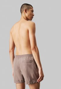 Pantaloni da nuoto color marrone chiaro, con vita elastica e una tasca posteriore con zip. Tessuto leggero con una texture liscia.