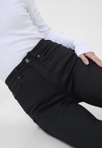 Jean noir ajusté en tissu extensible, avec une taille haute, des poches avant et une fermeture par bouton. Associé à un haut à manches longues blanc.