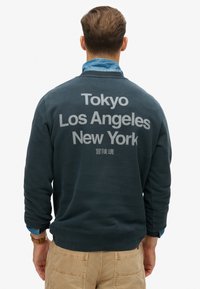 Sudadera de color verde oscuro con texto grande en gris: "Tokio Los Ángeles Nueva York" en la parte de atrás, con puños y dobladillo acanalados.