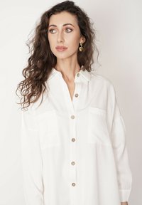 Biała koszula zapinana na guziki, wykonana z lekkiego materiału, z kołnierzykiem, dwiema kieszeniami na piersiach i brązowymi guzikami. Skóra gładka, a włosy kręcone.