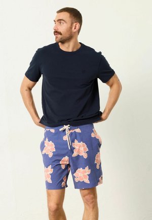 Bărbat cu mustață, purtând tricou negru și pantaloni scurți albaștri cu imprimeu mare floral roz, stând cu mâinile pe șolduri și uitându-se spre stânga.