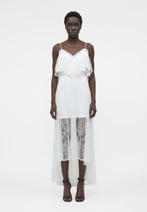 Γυναίκα με λευκό δαντελένιο φόρεμα με λεπτές τιράντες, στρωμένο μπούστο, διάφανο φούστα και μαύρα ψηλοτάκουνα με λουράκι στον αστράγαλο, σε απλό φόντο.