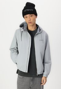 Lichtgrijze zip-up jacket met een capuchon, elastische manchetten en een matte afwerking. Draag het over een donker T-shirt en combineer het met grijze jeans en een zwarte beanie.
