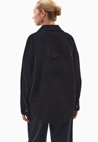 Czarna koszula z korduroju o luźnym kroju, z kołnierzykiem, z zapięciem na guziki przy mankietach oraz detale w tylnej części. Miękka tekstura z pionowymi prążkami.