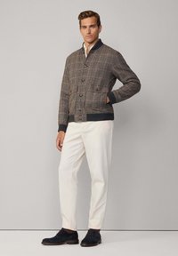 Chaqueta bomber a cuadros marrón con puños acanalados, bolsillos frontales y cierre de botón, combinada con pantalones blancos y zapatos oscuros.
