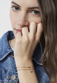Złota podwójna bransoletka z małą zieloną koraliką i wielokolorowym charmsiem w kształcie kwiatu, noszona na nagim nadgarstku na tle denimu.