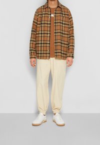 Camisa de franela a cuadros marrón y negro, camiseta marrón clara, pantalones crema ajustados, zapatillas blancas con suela de goma. Un atuendo simple y casual.