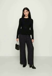 Μαύρος ραμμένος πλεκτός σκούφος συνδυασμένος με φαρδιά μαύρα παντελόνια με κεντήματα από παγιέτες. Το μοντέλο κρατά μια μικρή μαύρη τσάντα χεριού και φορά ankle boots. Ουδέτερο φόντο.