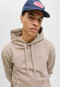 Sudadera de color beige con capucha y cordones ajustables, con bolsillo frontal, que presenta texto bordado en blanco. Combinada con una gorra de béisbol azul marino.