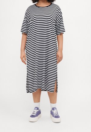 Persona con un vestido midi de rayas horizontales negras y blancas con aberturas laterales, calcetines blancos y zapatillas moradas, de pie contra un fondo blanco.