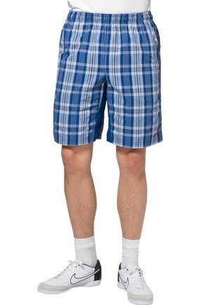 Blauwe geruite shorts met een elastische tailleband. Heeft afwisselend donkere en lichte blauwe vlakken. Draagt met witte sneakers en korte sokken.