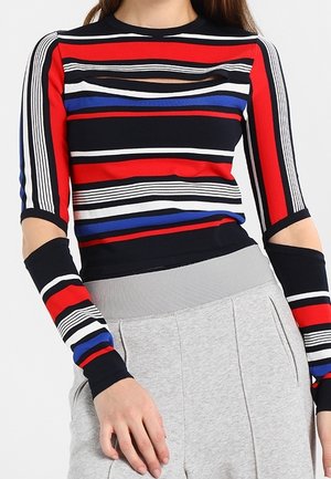Mujer con una blusa de manga larga a rayas de colores rojo, azul, negro y blanco, combinada con pantalones de talle alto en gris claro.