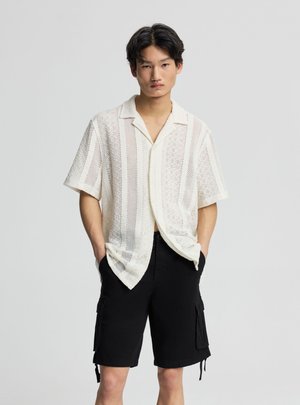 Giovane uomo con le mani in tasca, indossa una maglia bianca a maniche corte e pantaloncini cargo neri, in piedi davanti a uno sfondo chiaro e uniforme.