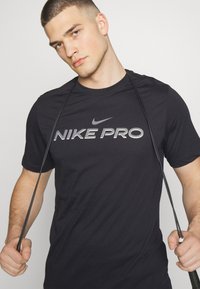 Must märg Nike Pro T-särk, mille esiküljel on suur "NIKE PRO" logo valge ja halliga. Lühikesed varrukad, ümmargune kaelus ja sile kangastekstuuri.