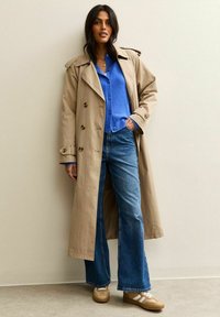 Beżowy płaszcz trench z podwójnymi guzikami, w połączeniu z niebieską koszulą i dżinsami z rozszerzanymi nogawkami. Beżowe trampki z białymi akcentami.