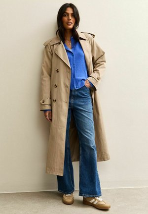 Beige trenchcoat met dubbele knopen, gecombineerd met een blauwe shirt en uitlopende denim jeans. Tan sneakers met witte accenten.