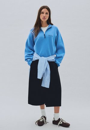 Junge Frau trägt einen hellblauen Viertel-Reißverschluss-Pullover, schwarzen Midirock, weiße Socken, braune Schuhe und ein blau gestreiftes Hemd, das um die Taille gebunden ist.