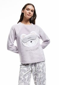 Camiseta de punto de color púrpura claro de manga larga con un rostro de oso y el texto "ESTA ES LA ZONA DE DESCANSO"; combinada con pantalones de pijama color crema con ilustraciones de osos.