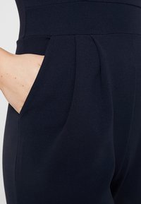 Main placée dans la poche latérale d'un pantalon sur mesure bleu marine foncé avec une taille haute et un détail plissé à l'avant.