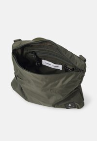 Borsa in tessuto leggero verde oliva con zip superiore e tasca interna. Presenta un'etichetta del marchio e una superficie texturizzata.