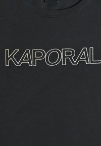 Czarna bawełniana koszulka z wyrazem "KAPORAL" w pogrubionym, białym konturowym druku, umieszczonym centralnie na piersi.