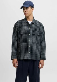 Camicia azzurra a quadri con due grandi tasche sul petto, chiusura a bottoni e una vestibilità comoda, indossata sopra una maglietta bianca e abbinata a pantaloni scuri.