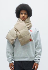 Bufanda acolchada beige hecha de tela suave, envuelta alrededor del cuello. Etiqueta de logo roja visible. Modelo luciendo un sudadera gris claro y pantalones oscuros.