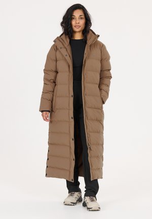 Kvinde iført en lang brun polstret jakke med hætte, sorte bukser og lyse sneakers, stående mod en ensfarvet baggrund.