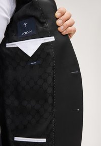 Interno di una giacca nera con fodera decorata, dettaglio bianco sulla tasca e etichette per marca e vestibilità. Tessuto strutturato con cuciture ordinate.