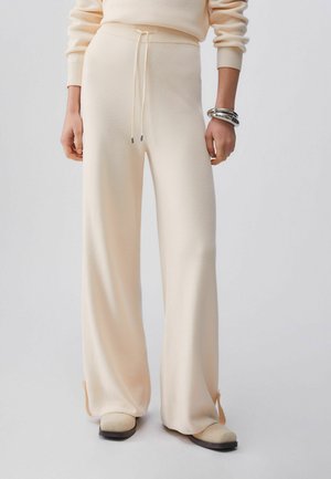 Cremefarbene, weit geschnittene Strickhose mit Kordelzug in der Taille, getragen mit passendem Oberteil und beigen Schuhen, silberne Armbänder am linken Handgelenk.