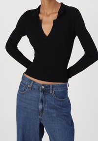 Černý žebrovaný top s dlouhým rukávem, V-výstřihem a límečkem, v kombinaci s světle modrými džíny s vysokým pasem, které mají knoflíkové zapínání a kapsy.