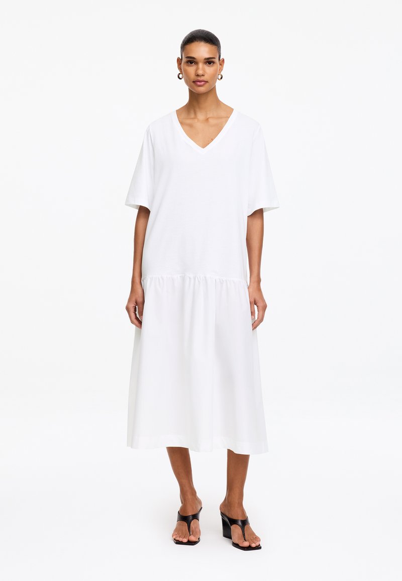 Femme aux cheveux foncés portant une robe midi blanche ample à col en V et des sandales noires à talons, debout devant un fond blanc uni.
