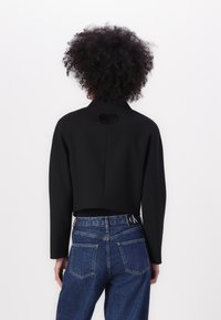 Zwarte cropped jas met een kraag, lange mouwen en een label aan de achterkant. Gecombineerd met blauwe hoge taille denim jeans met zichtbare stiksels.