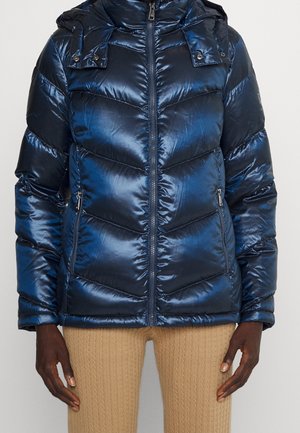 Doudoune bleu marine brillante avec fermeture éclair frontale et poches zippées portée avec un pantalon en maille beige sur une personne à la peau foncée.
