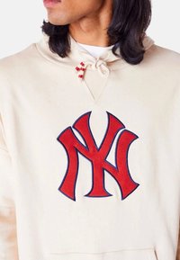 Felpa beige con un logo dei New York Yankees ricamato in rosso ben visibile. Collo regolabile con un cordino crema, tessuto morbido.