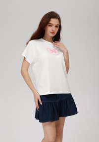 Camiseta de algodón blanca con mangas cortas, que presenta un diseño de corazón rosa con un lazo en el centro. Combinada con una falda de mezclilla oscura con volantes.