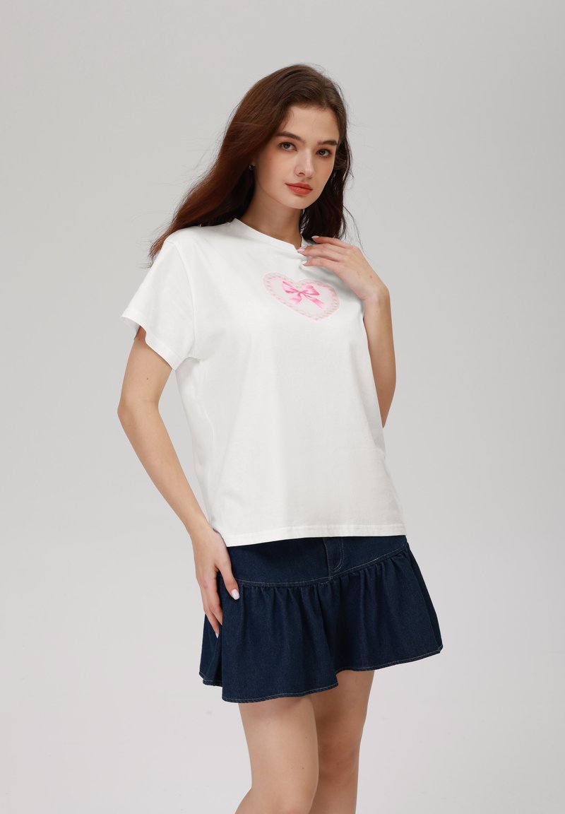 Camiseta de algodón blanca con mangas cortas, que presenta un diseño de corazón rosa con un lazo en el centro. Combinada con una falda de mezclilla oscura con volantes.