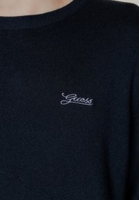 Maglione a maglia blu navy con scollo rotondo, caratterizzato da un piccolo logo ricamato bianco "Guess" nella zona sinistra del petto.