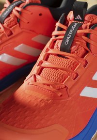 Orange athletic shoes with mesh and synthetic upper, featuring black tongue label, white stripes, and textured surface. Blue sole base.