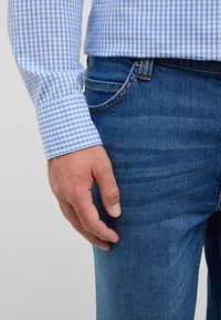 Jasnoniebieska koszula w kratkę z białymi elementami i mankietami na guziki, zestawiona z niebieskimi dżinsami. Widoczna ręka oparta na dżinsach.