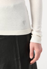 Pull en tricot couleur crème avec des manches longues, présentant une finition texturée et un petit logo triangulaire en strass sur le côté inférieur. Porté avec une jupe en cuir noire.