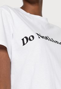 Vit bomull t-shirt med kort ärm; har svart broderad text "Do Nothing" på framsidan, slät textur och rundad halsdesign.
