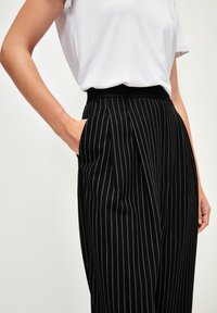 Pantalons noirs à rayures avec un design plissé, taille élastique et poches latérales. Le tissu a une texture lisse.